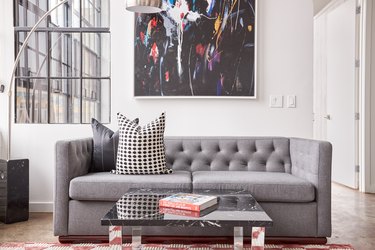 Modern living room with gray couch, mirror coffee table, large metal floor lamp, contemporary art, black, white and gray pillows.